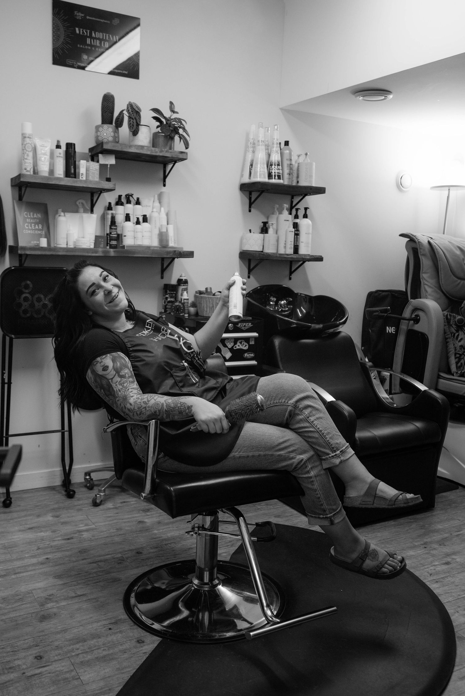 Black and white photo of tattooed female hairstylist relaxing in salon styling chair laughing.
