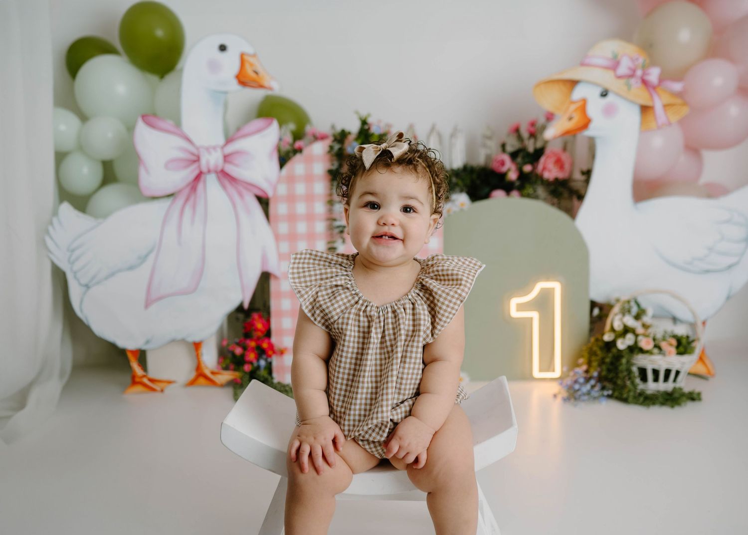 A baby girl in a gingham romper sits smiling at her first birthday cake smash with goose balloons and a glowing number one.
