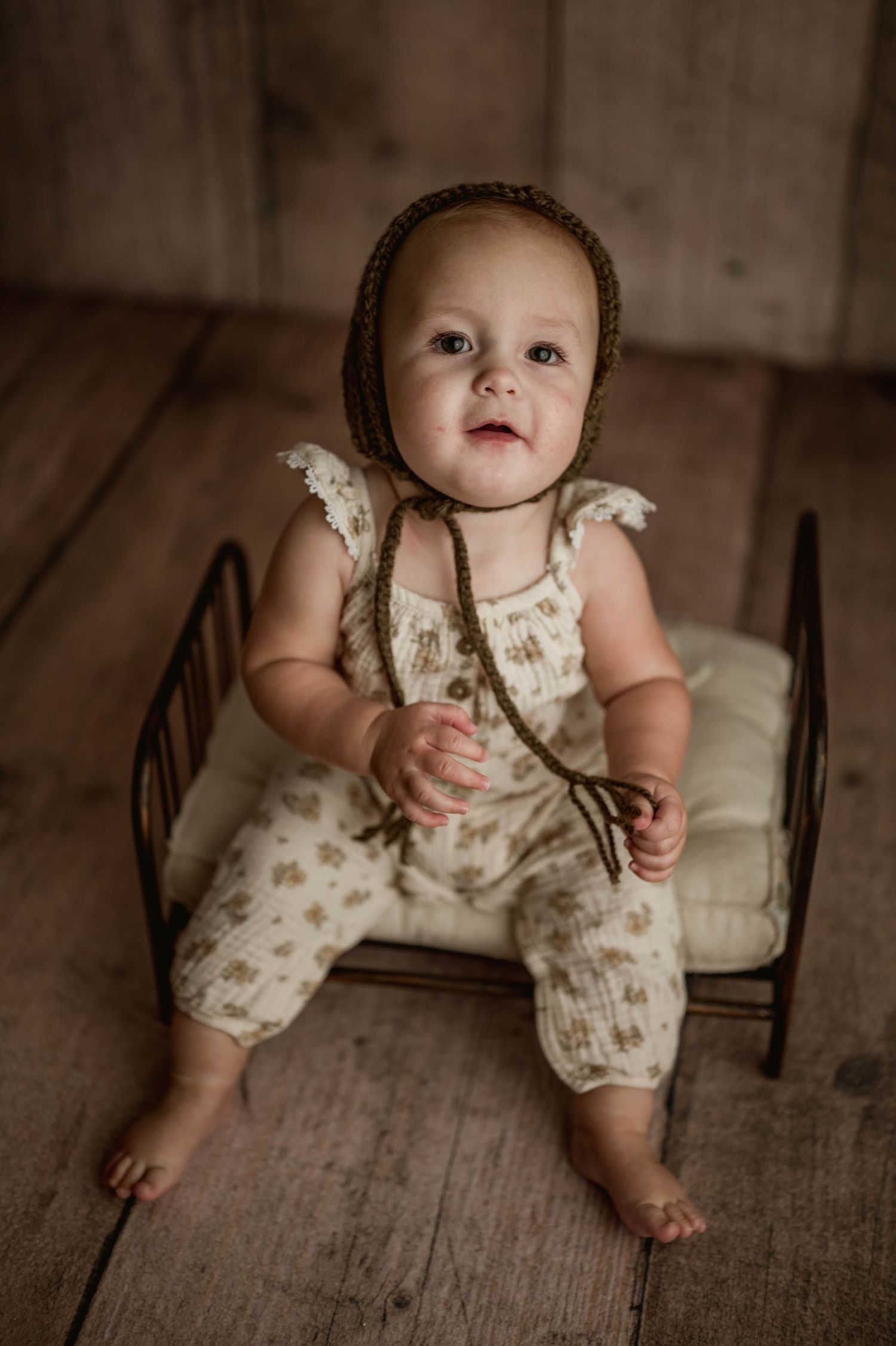 Baby in floral romper and brown knit bonnet sits on miniature iron bed during studio portrait.