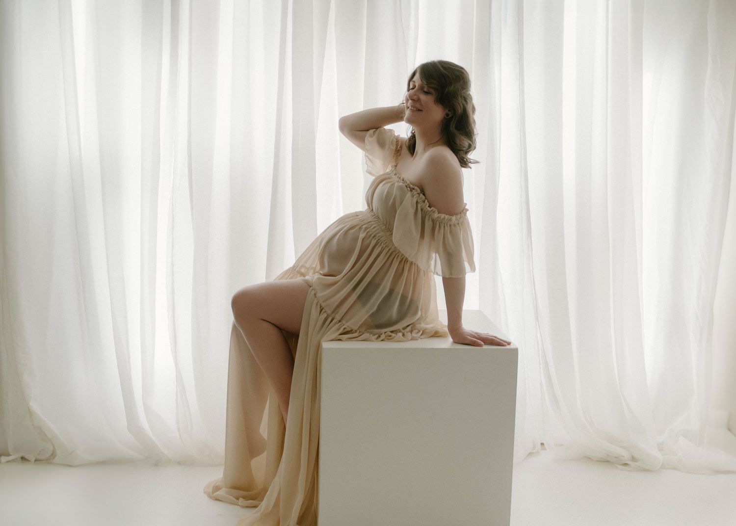 Pregnant woman in sheer gold gown sitting gracefully on white cube against flowing white curtains in a Leduc Photo studio.