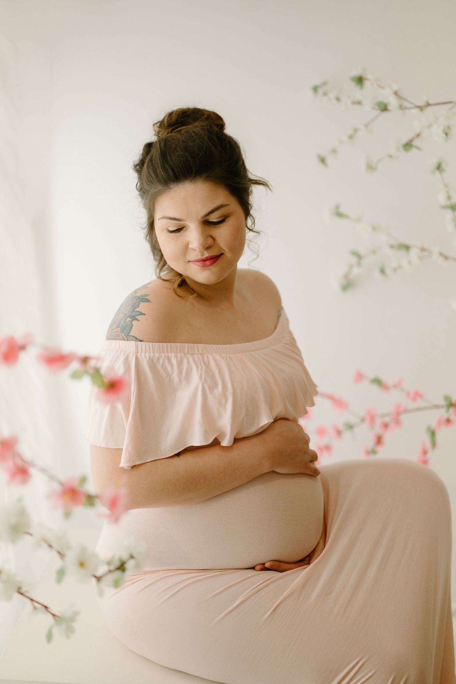 Pregnant woman with shoulder tattoo in blush off-shoulder ruffle dress tenderly cradles baby bump near blossoms.