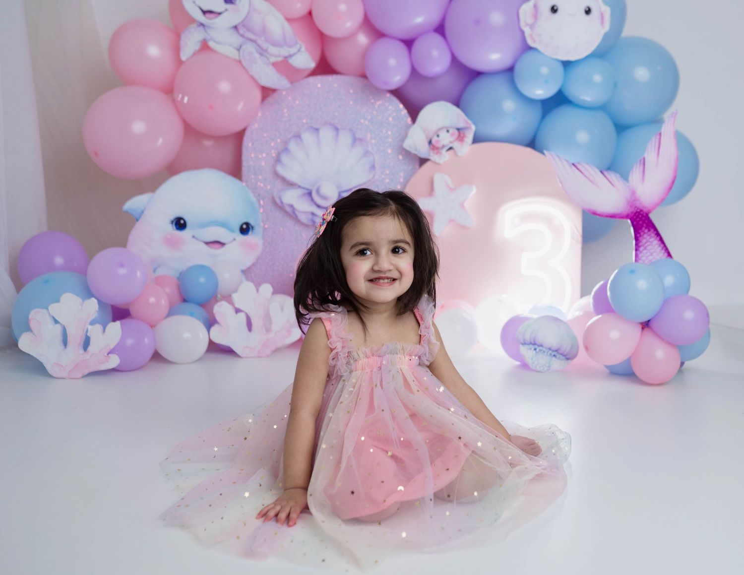 Happy little girl in sparkly pink dress posing at mermaid themed third birthday party setup.