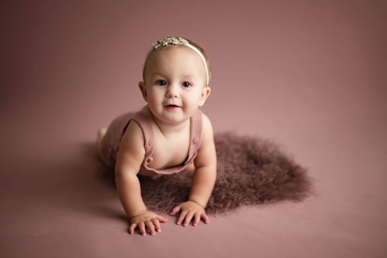 Baby girl in pink romper and floral headband crawling on fluffy brown rug against mauve background.