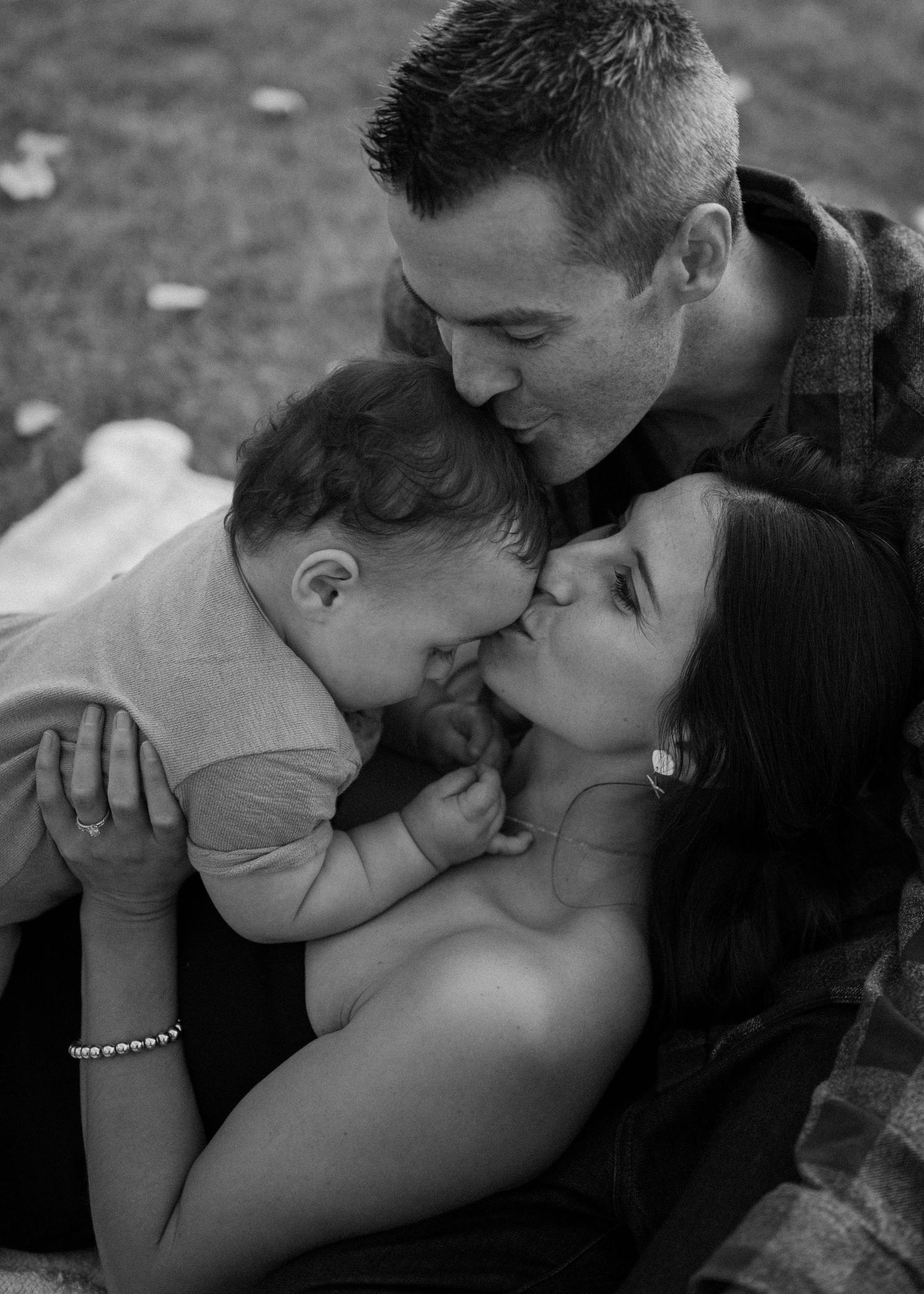 Black and white photo of loving parents kissing their smiling baby together outdoors.