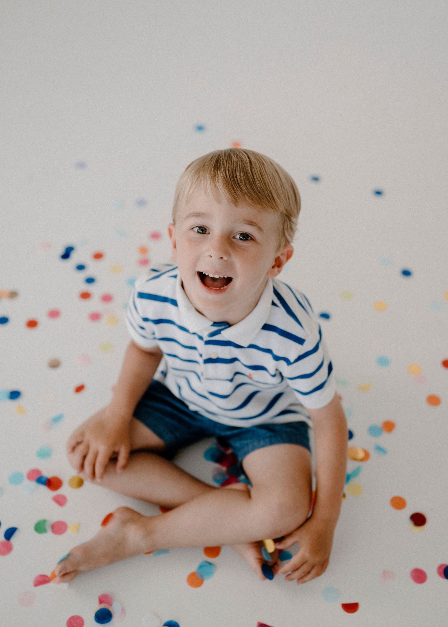Happy blonde boy sitting on white floor surrounded by colorful confetti, laughing joyfully.