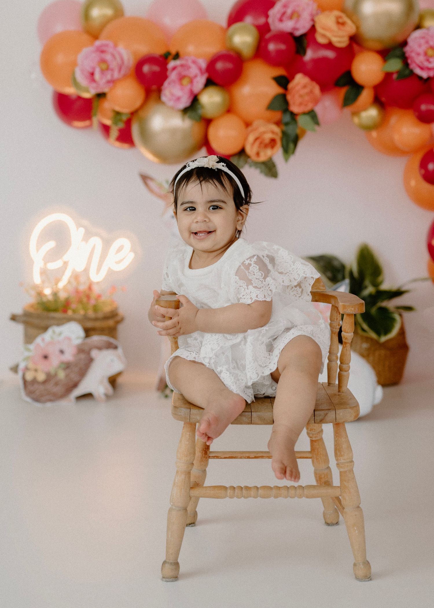 Happy baby girl in white lace romper sitting on wooden chair at first birthday celebration.