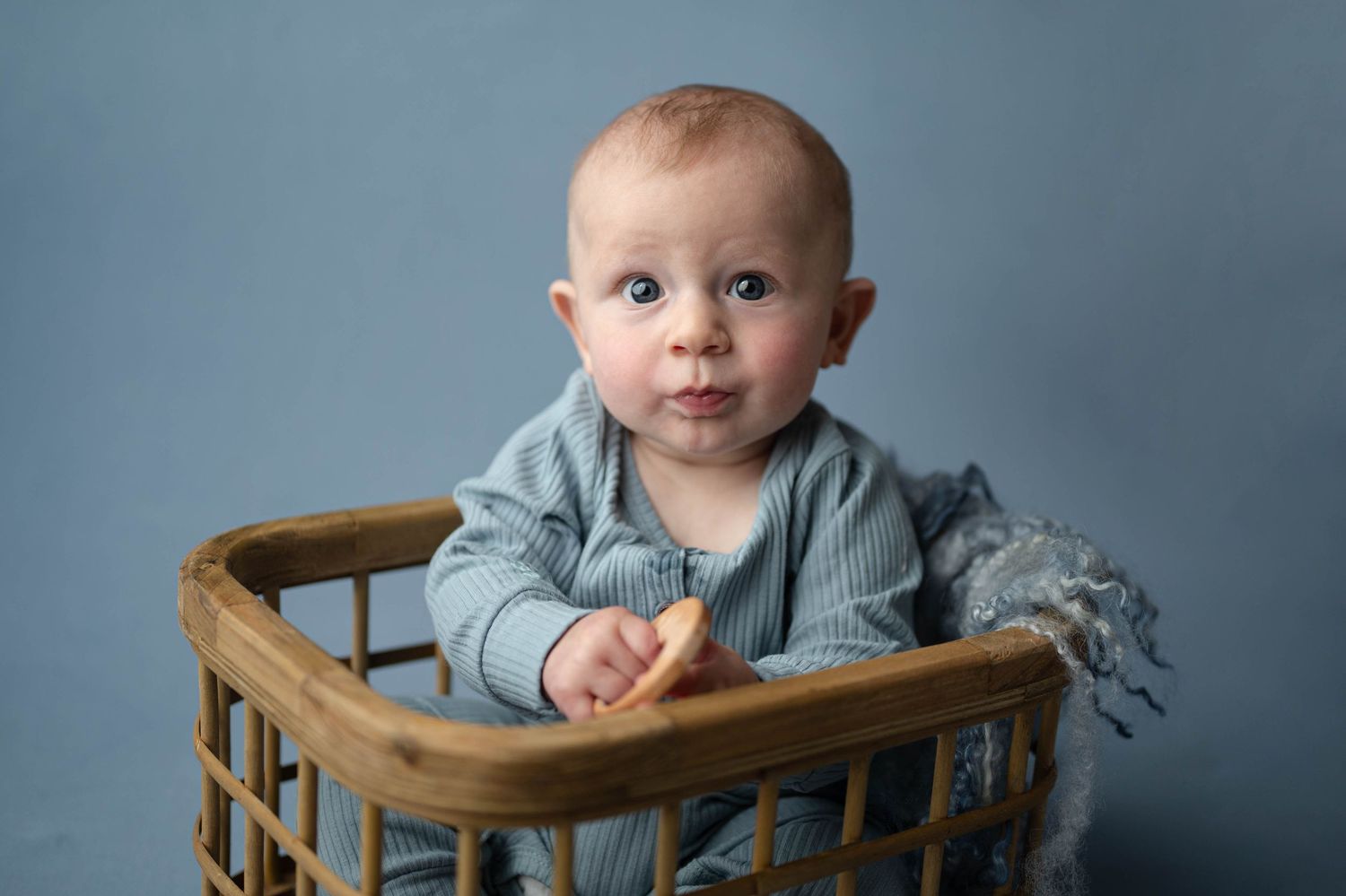 Cute baby in a striped blue outfit sitting in a wicker basket on a blue background.