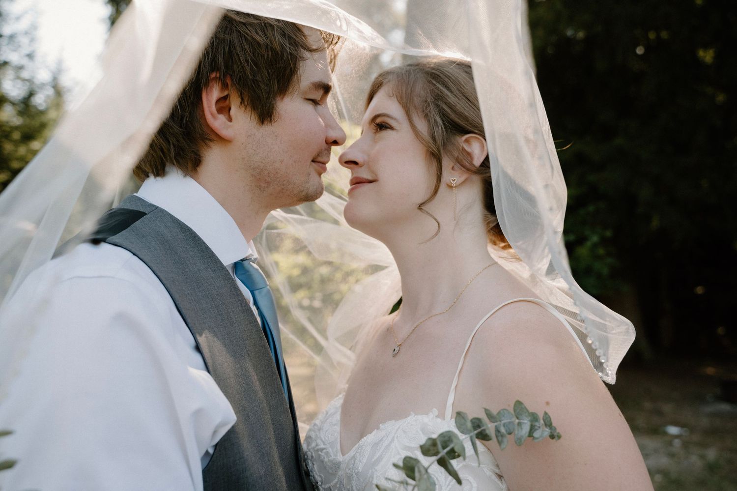 Bride and groom share a romantic moment under a flowing veil in a sun-dappled outdoor setting.