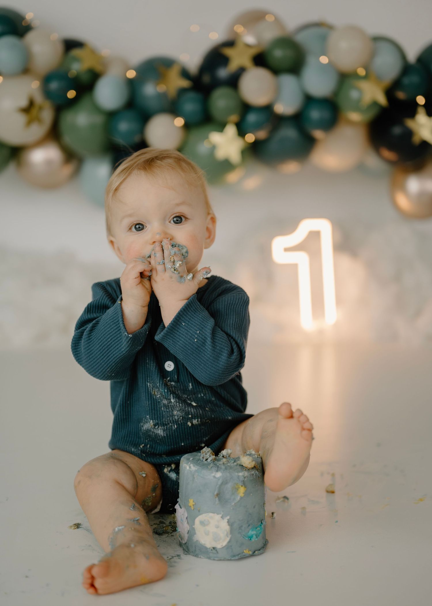 Baby boy in teal romper smashing first birthday cake with balloon garland and neon number 1.