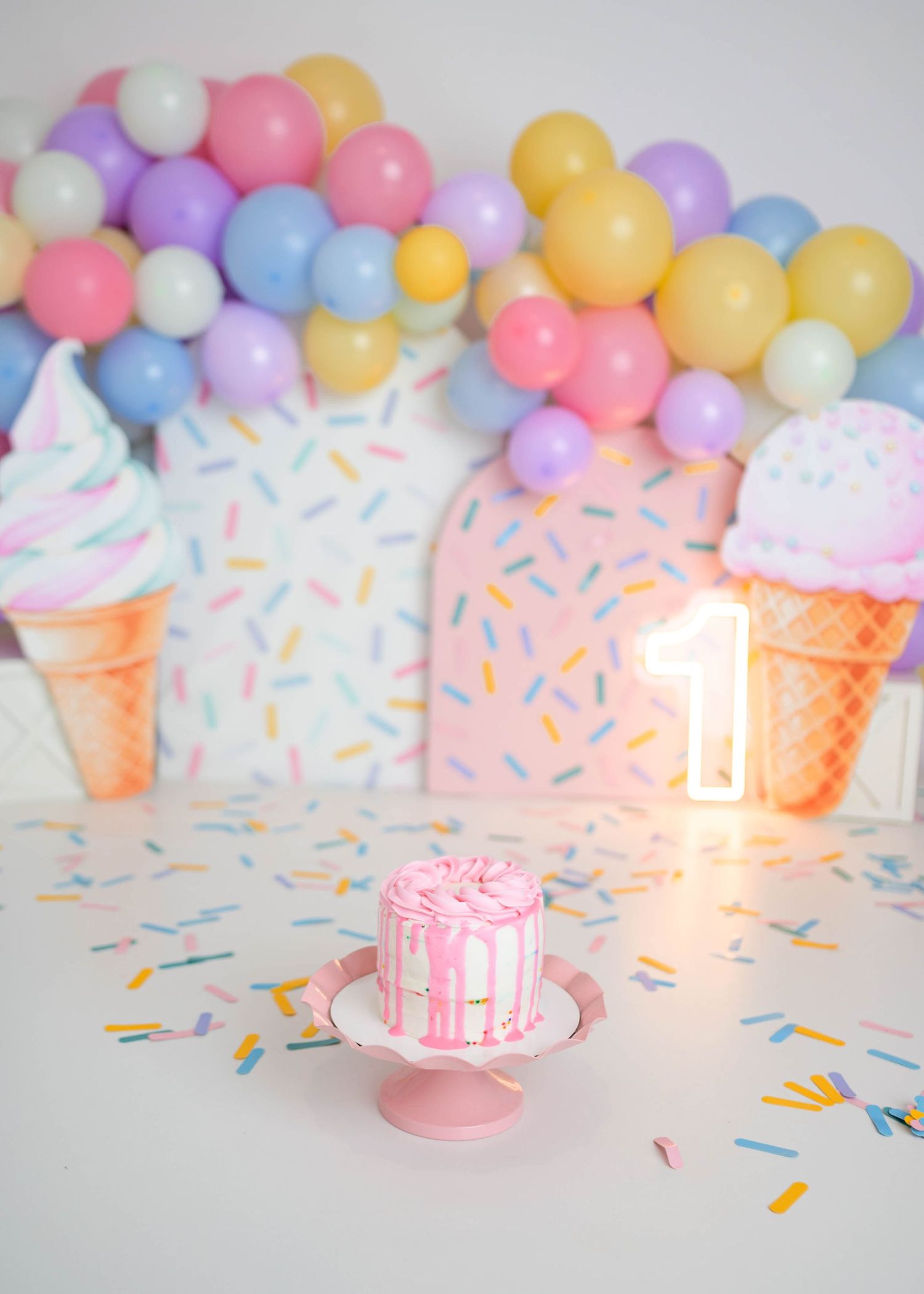 Pink drip smash cake on stand at ice cream themed first birthday party with colorful balloon backdrop in a leduc photo studio.