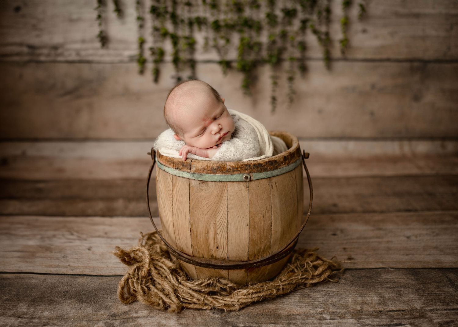 Sleeping newborn baby curled up in a rustic wooden barrel bucket on a wood plank floor.