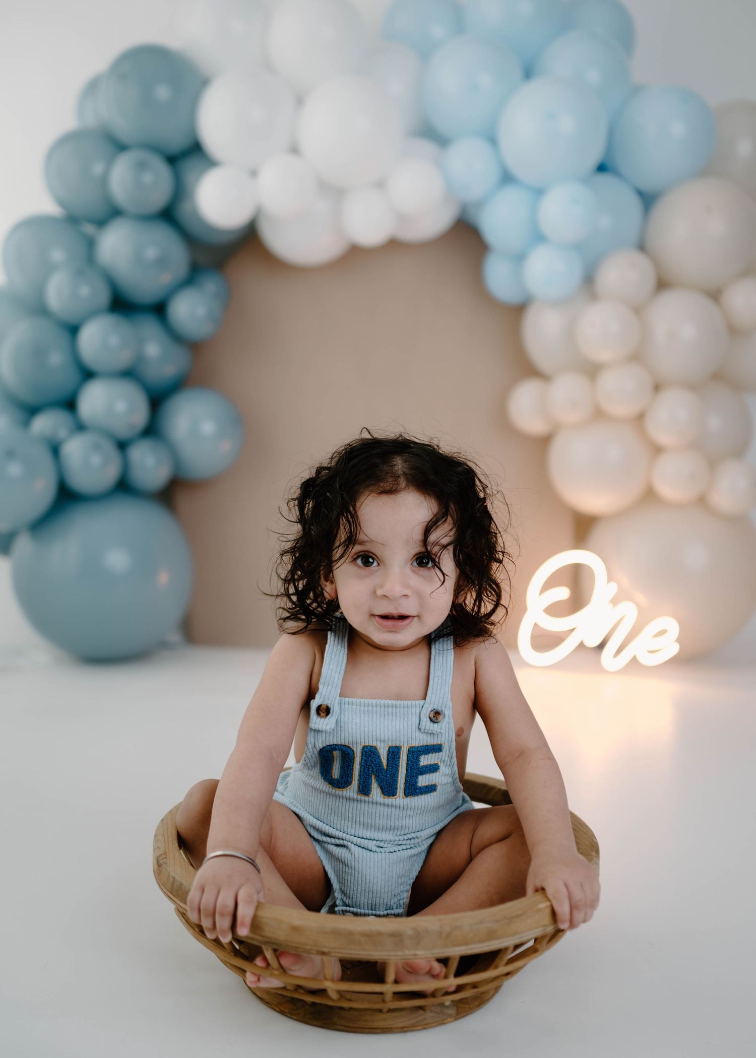 Cute toddler wearing ONE romper sits in wicker basket in front of blue and white balloon arch with neon sign.
