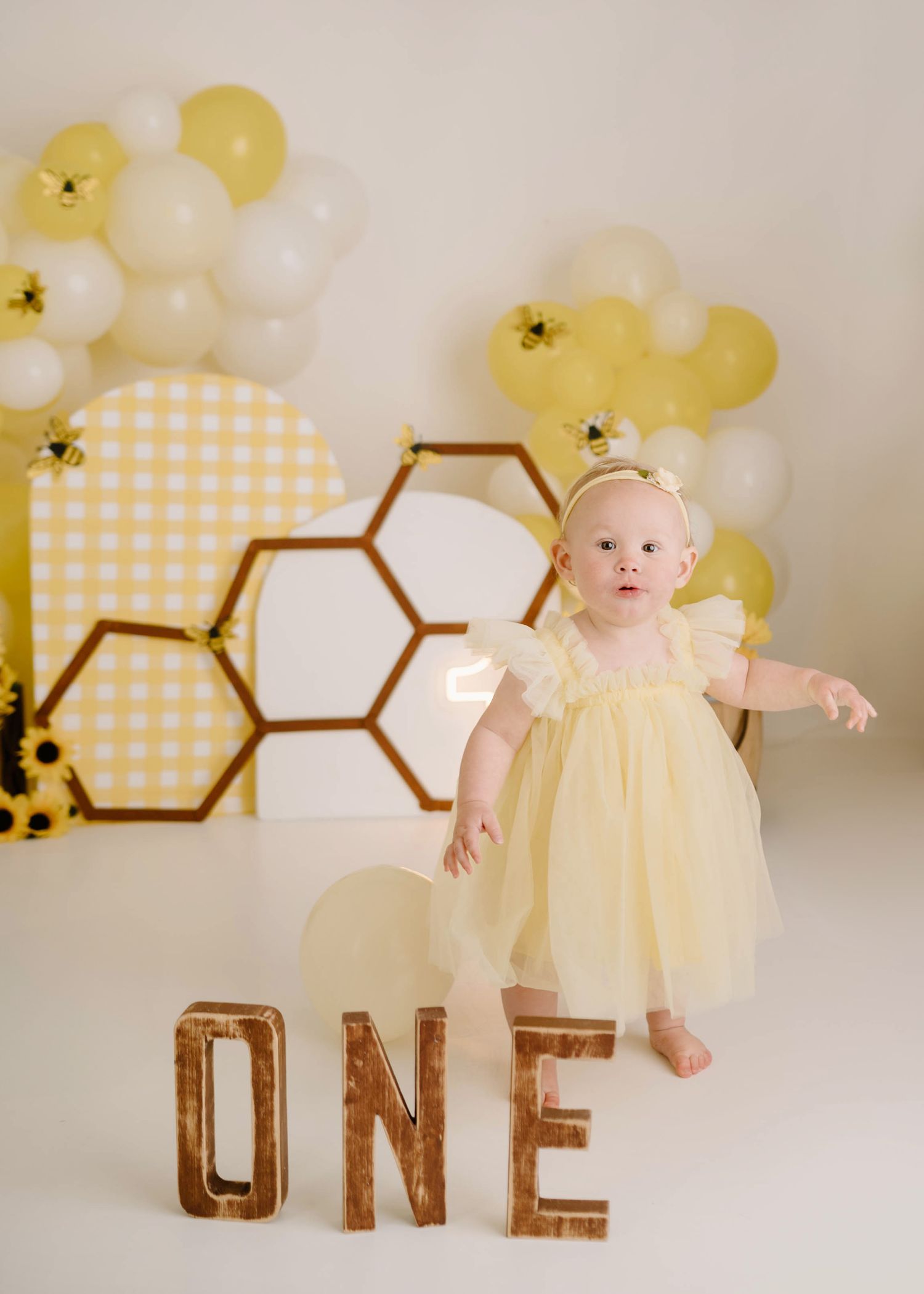Baby girl in yellow tulle dress stands by ONE letters at bee-themed first birthday party with balloon arch.