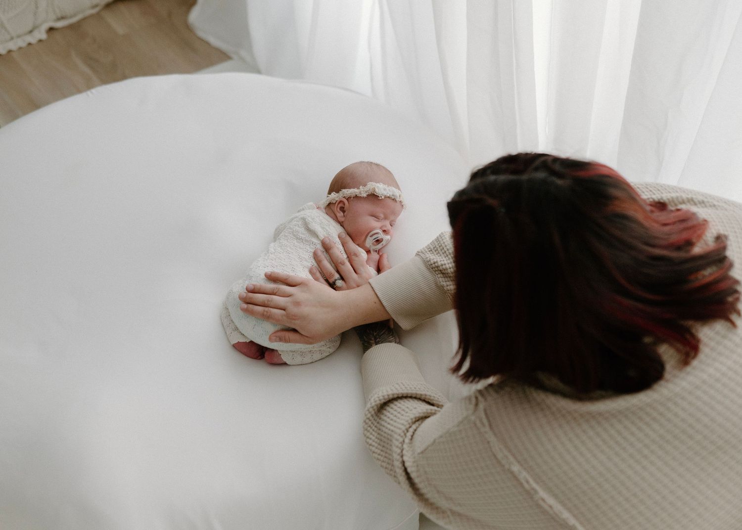 A mother tenderly gazes at her swaddled newborn baby lying on a white round bed in a bright, airy room.