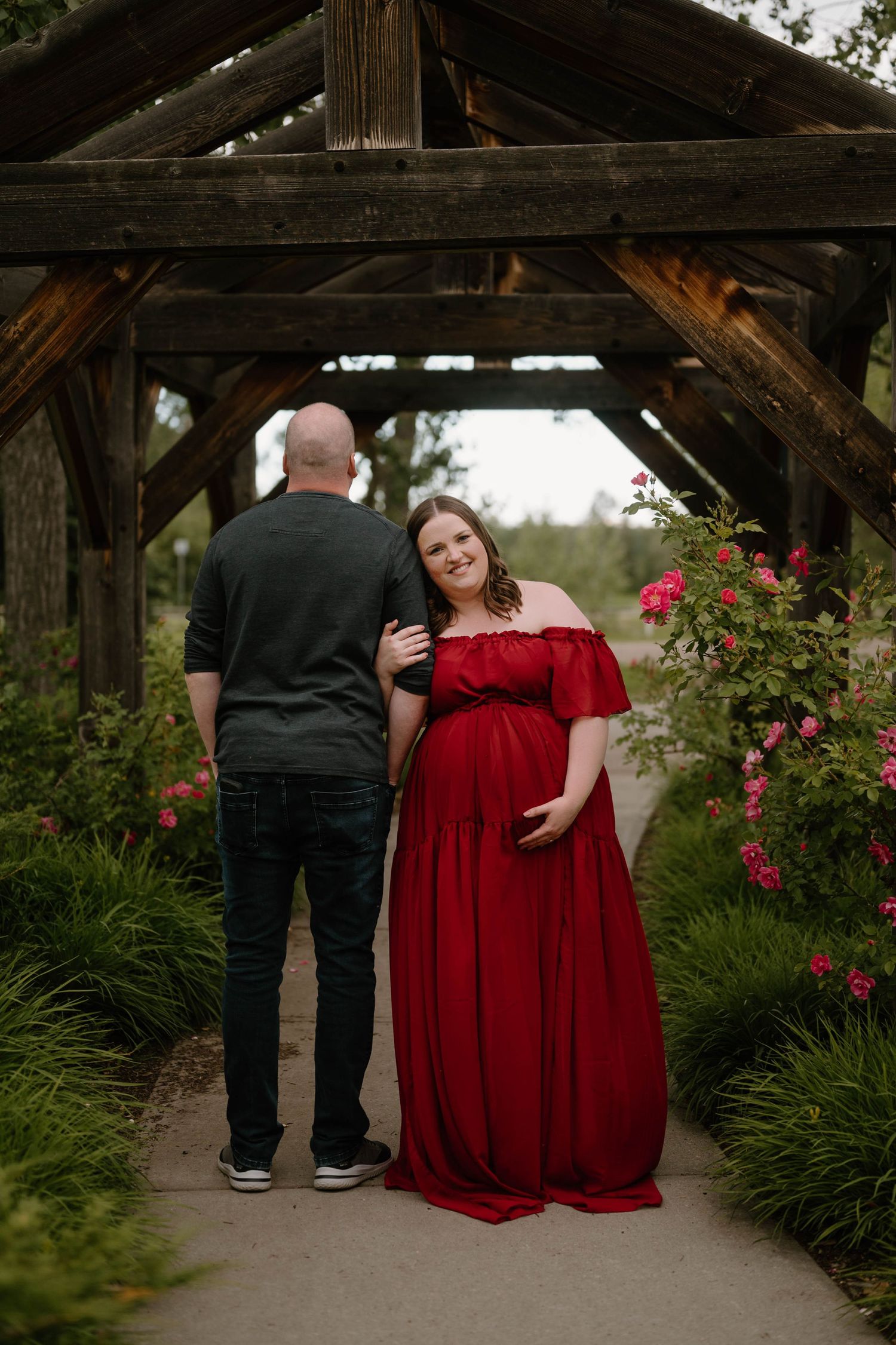 Pregnant woman in red off-shoulder gown poses with partner under wooden arbor surrounded by blooming roses.