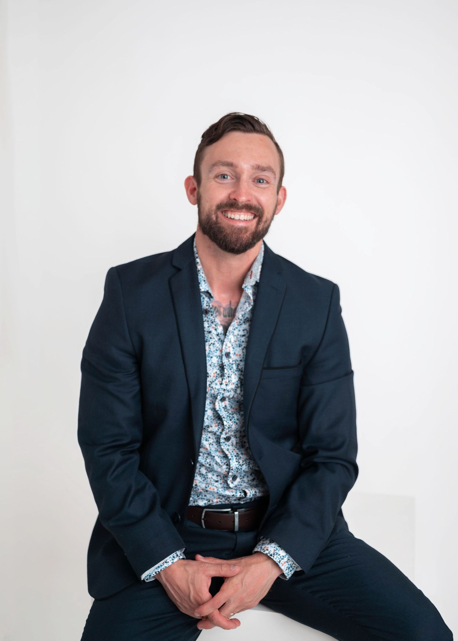 Smiling bearded man in navy blazer and floral shirt sits confidently against white background for professional headshot.