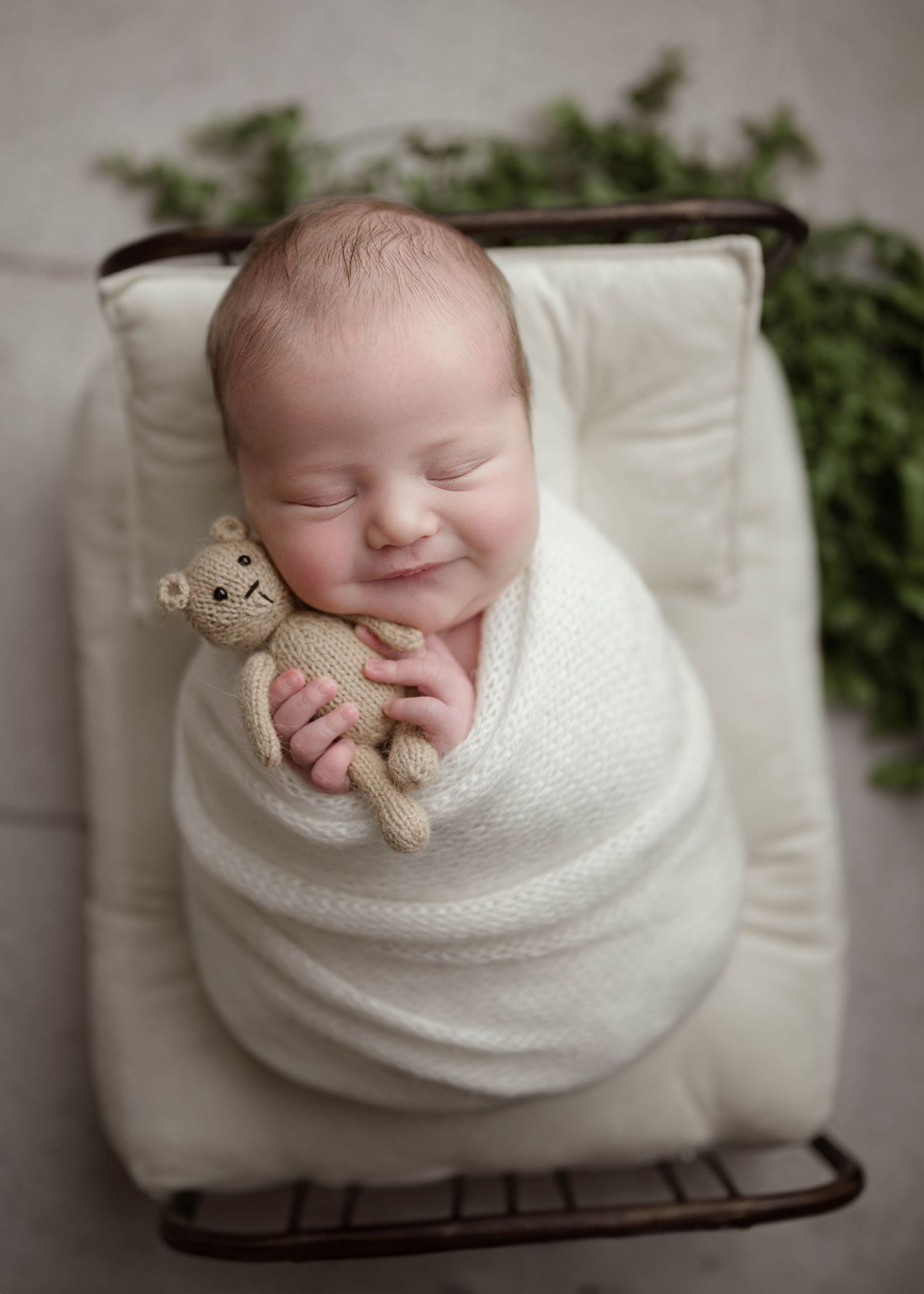 Sleeping newborn baby swaddled in white wrap holding a knit teddy bear in a vintage metal bed.