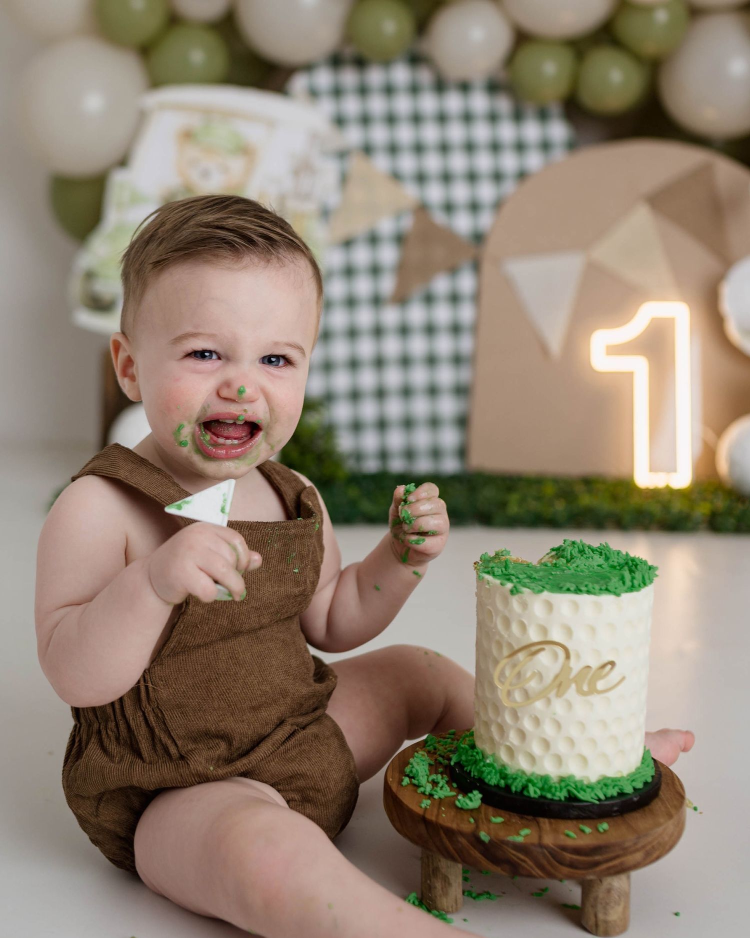 Smiling baby boy celebrating first birthday with golf-themed cake smash in brown romper with green frosting on face.