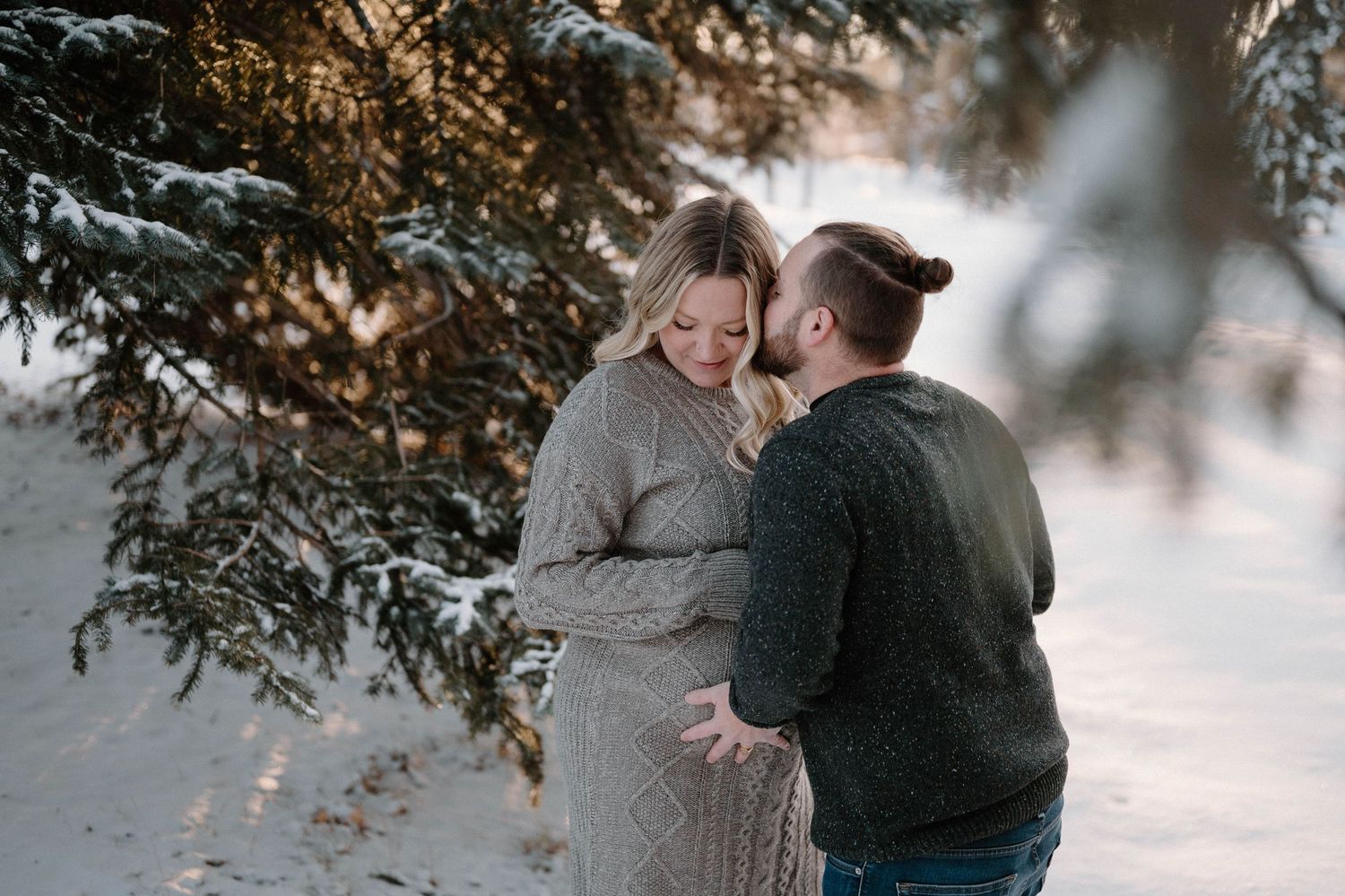 Expecting couple embracing under snowy pine trees during golden hour winter maternity session.