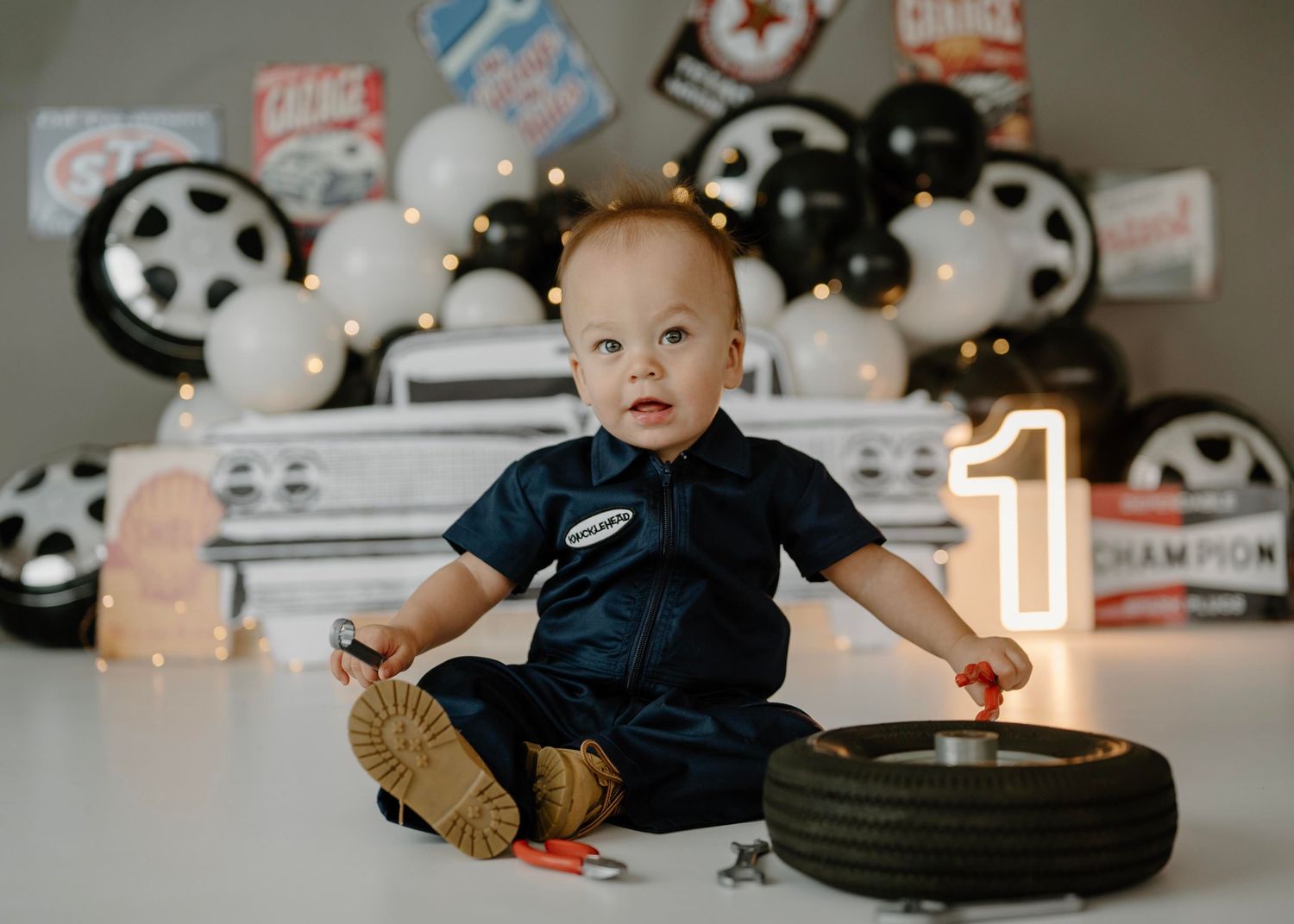 One year old boy in mechanic costume plays with tire and tools at garage birthday cake smash shoot.