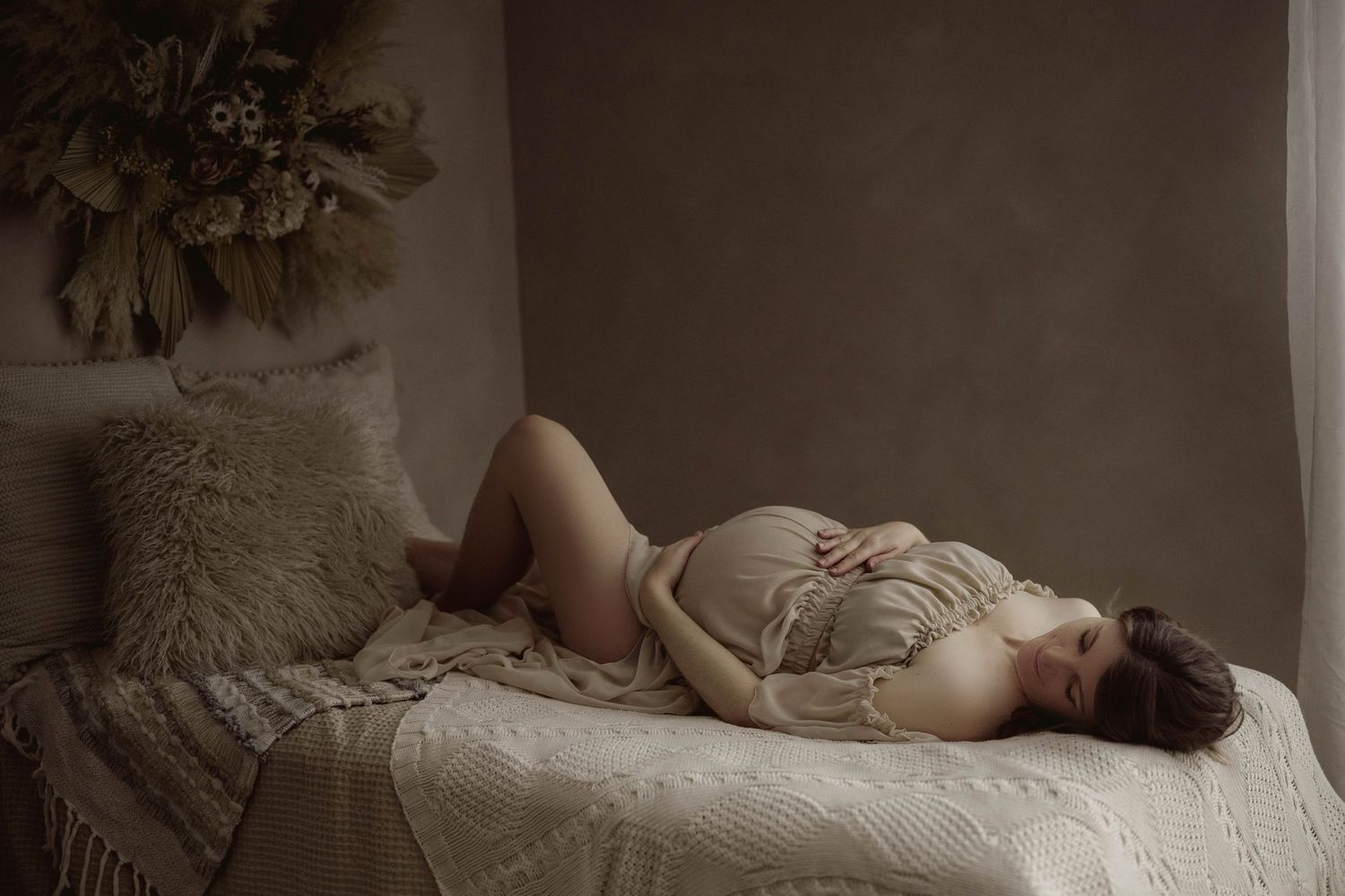 Pregnant woman resting on a bed in a draped wrap, posing in a moody, warmly lit studio setting.