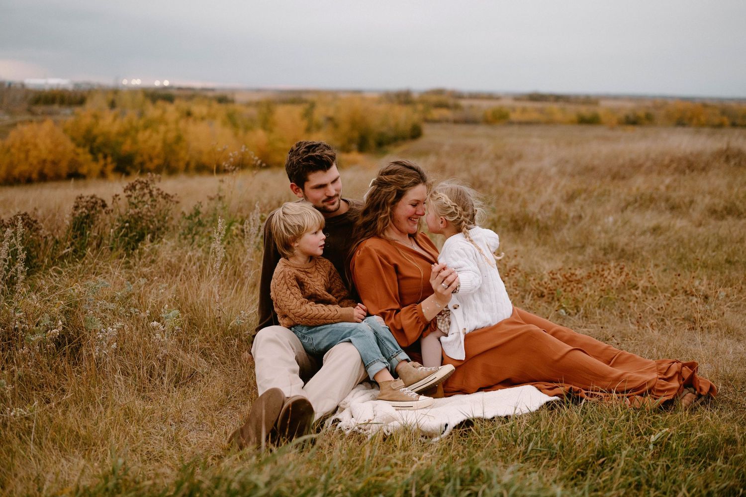 Happy family of four sitting together on a blanket in an autumn field, laughing and embracing.
