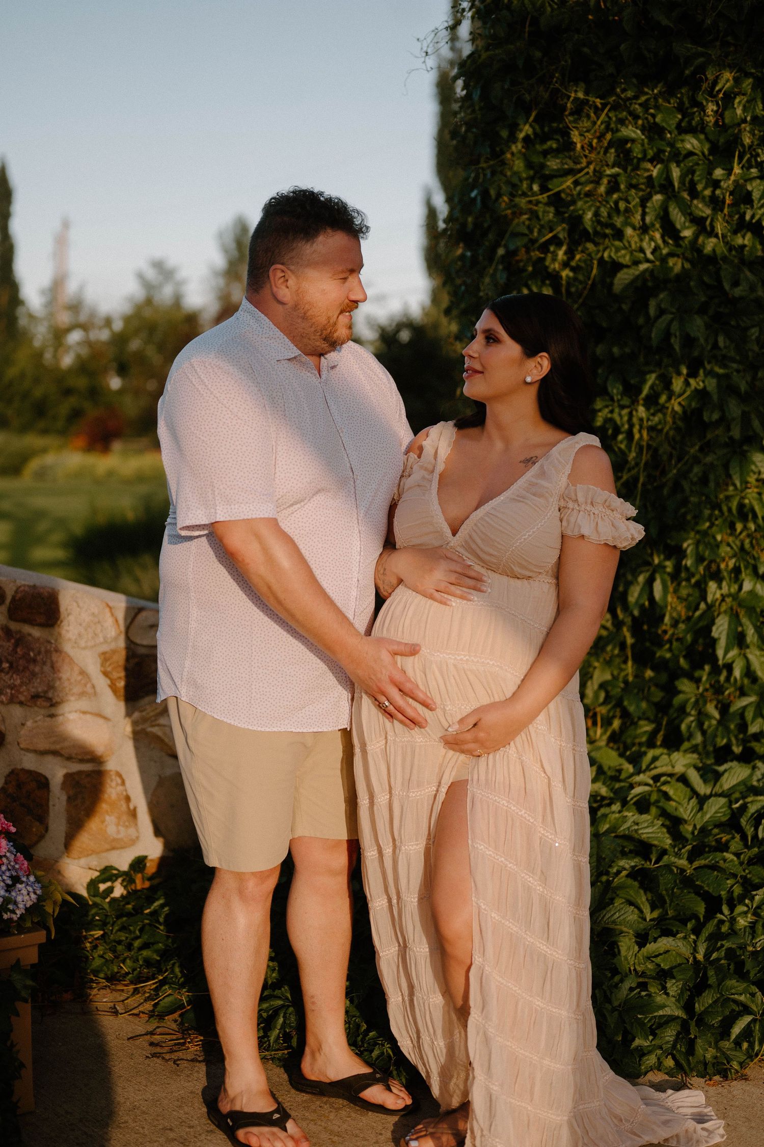 Couple sharing a tender moment outdoors, woman in flowing maternity dress cradling baby bump at golden hour.