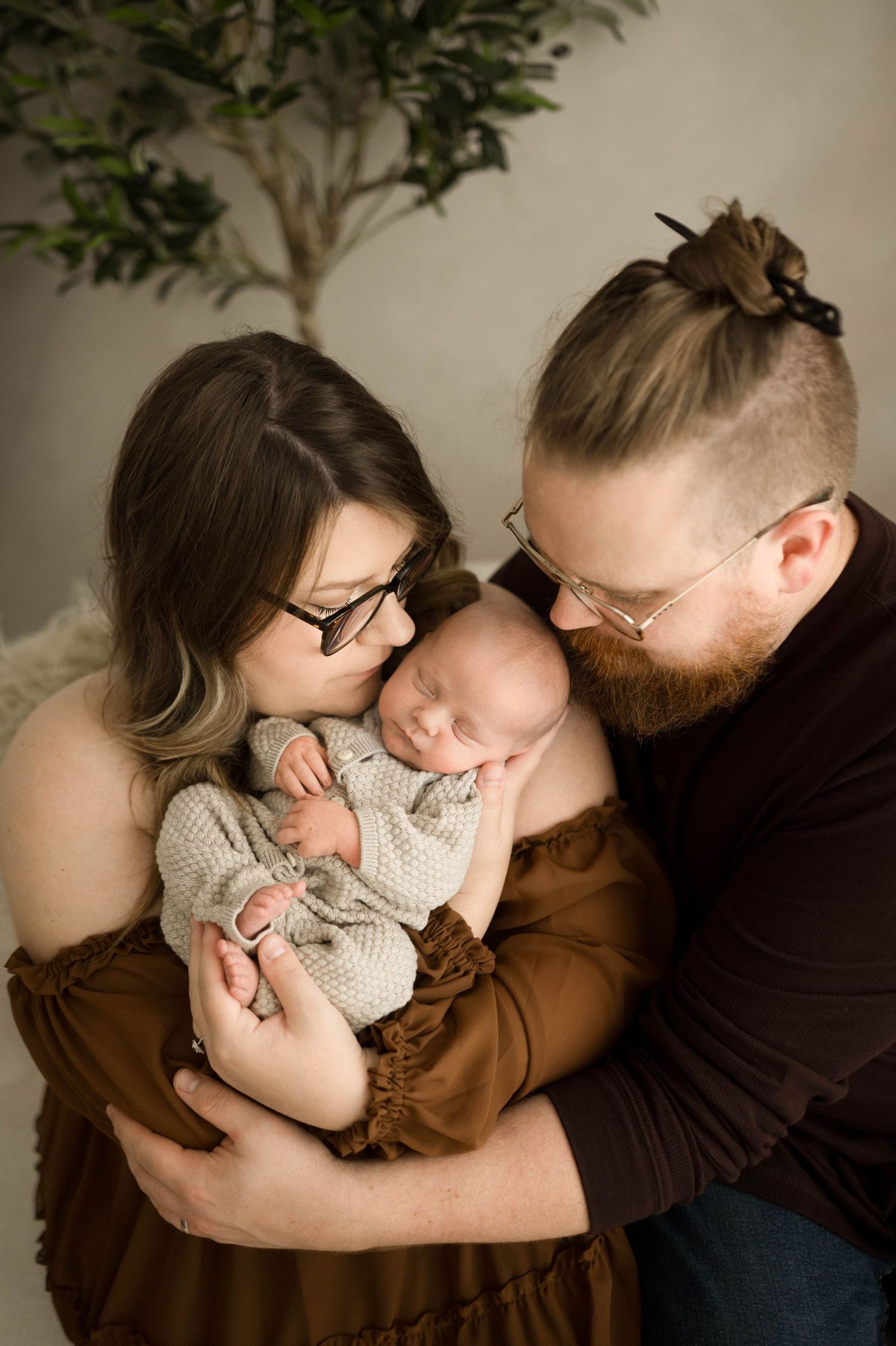 New parents in brown outfits tenderly kiss their sleeping newborn baby during a professional studio portrait session.