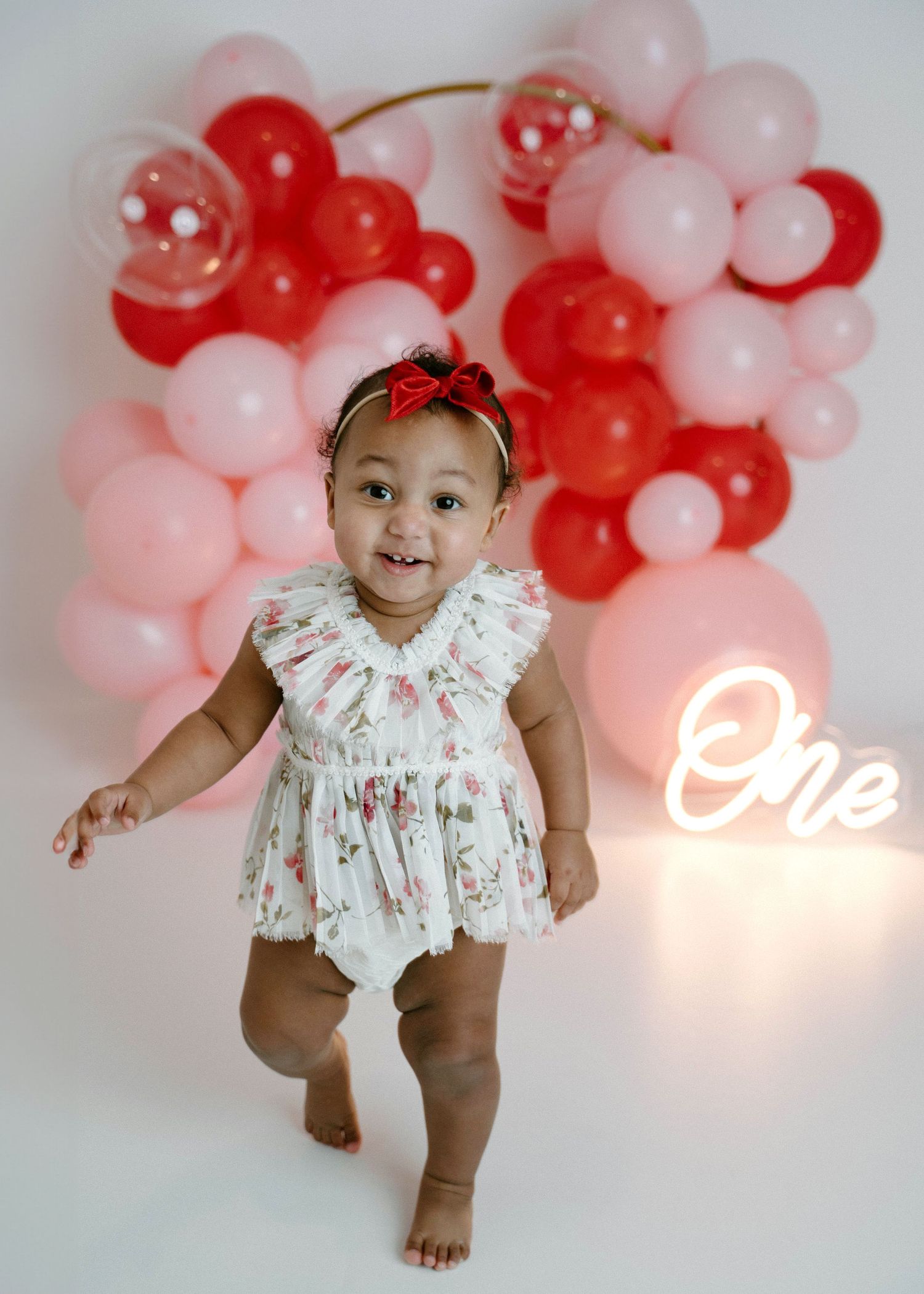 Smiling baby girl stands in floral outfit with red bow headband before pink red balloon display.