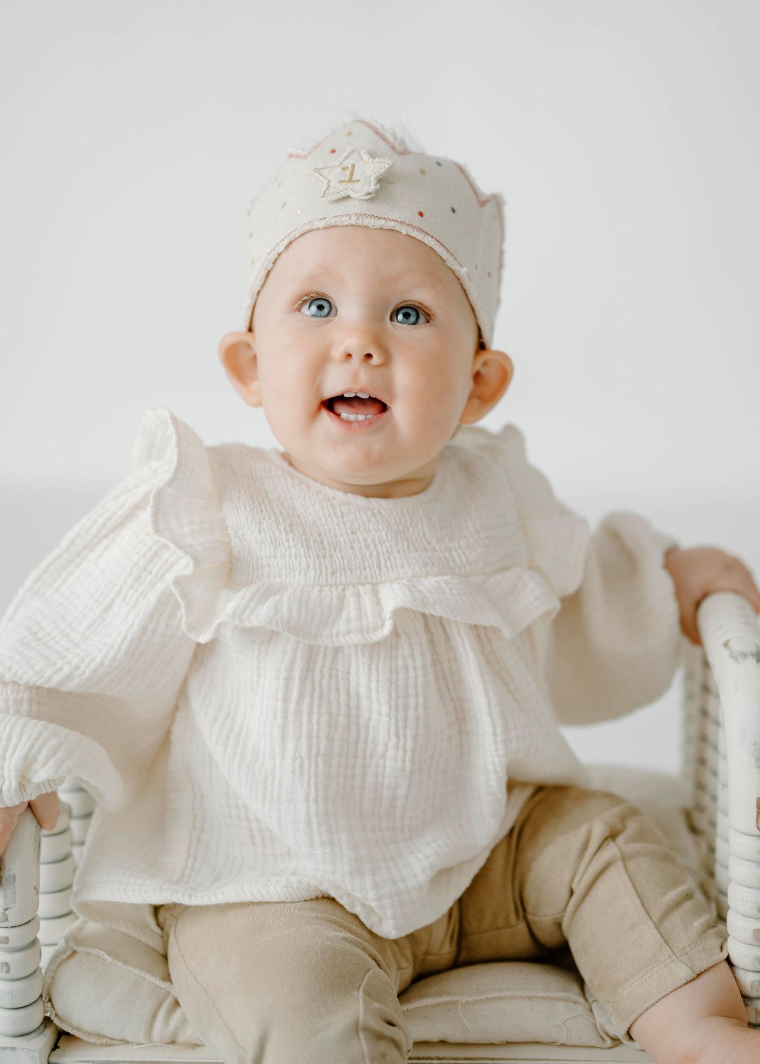 Blue-eyed baby in white ruffle top and crown sitting in white wicker chair smiling.