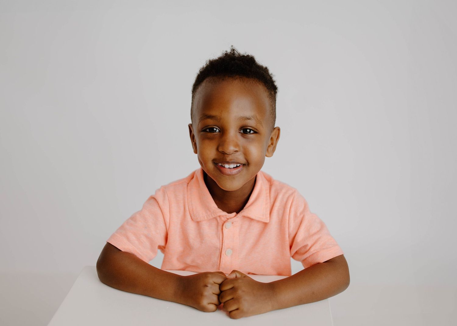 Smiling young Black boy in a pink polo shirt sitting at a white table against a light gray background.