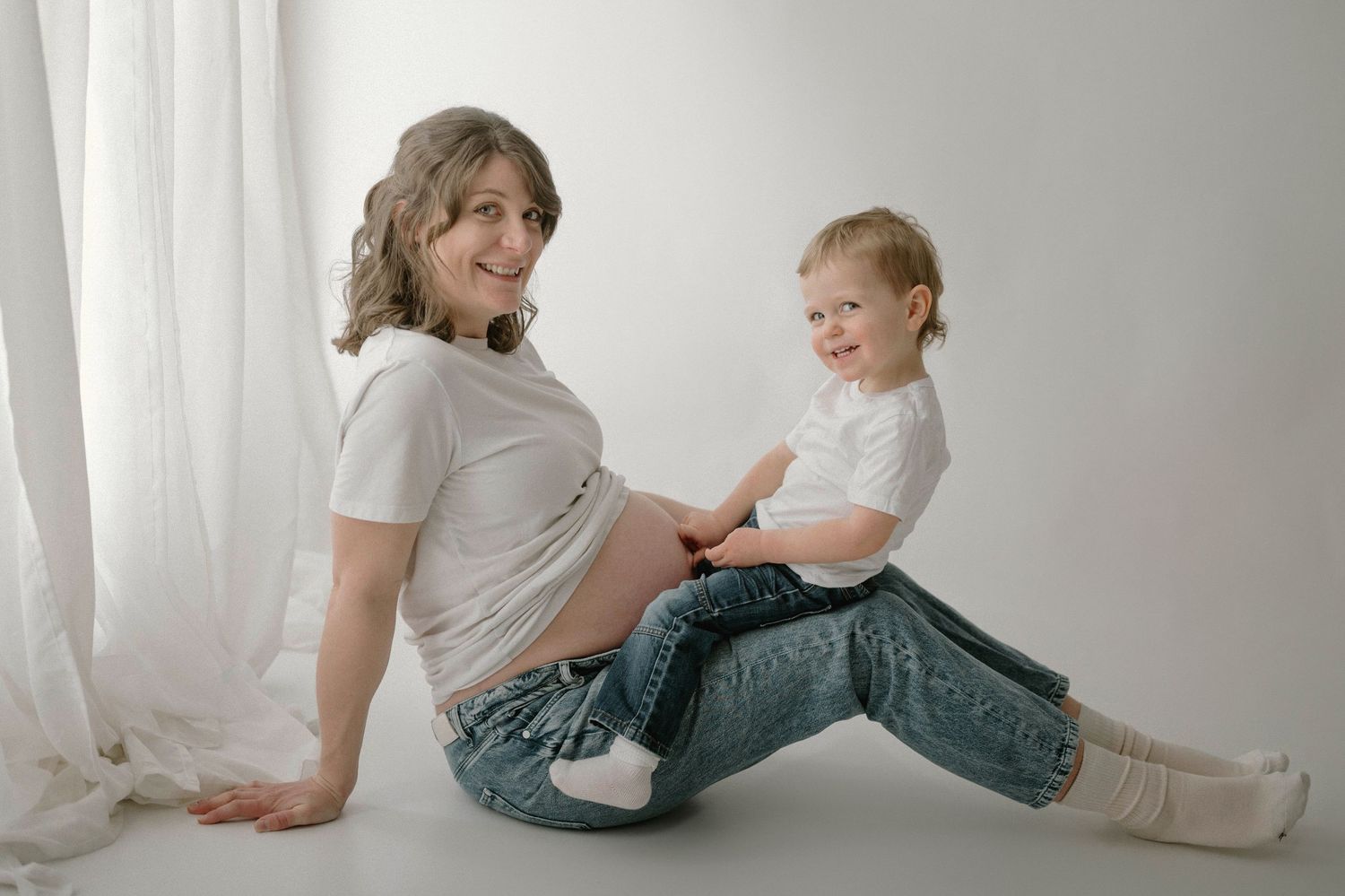 Smiling pregnant woman in white tee and jeans sitting on floor with her toddler in a bright studio.