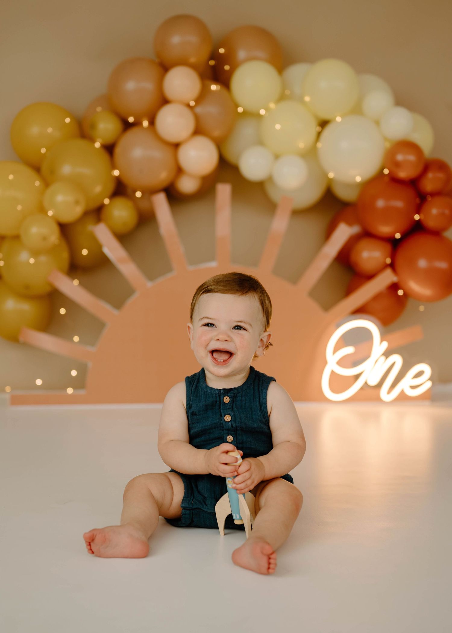 Happy laughing baby boy in teal romper with boho rainbow balloon arch and One neon sign.