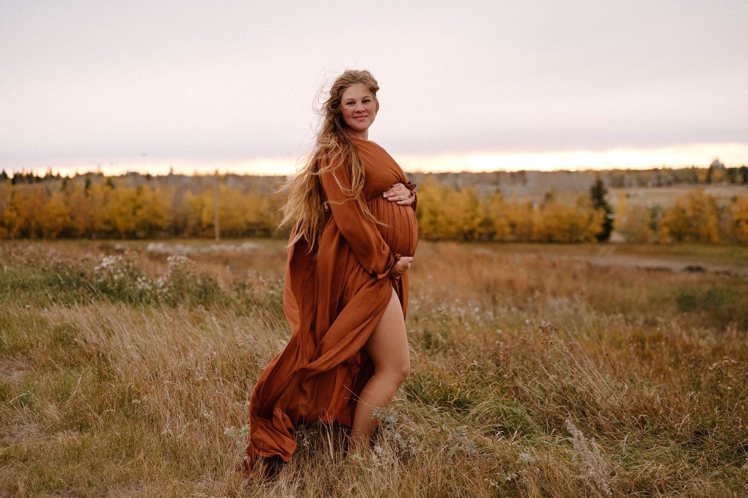 Pregnant woman in flowing rust-colored maternity gown posing in autumn field In Leduc Alberta at sunset.