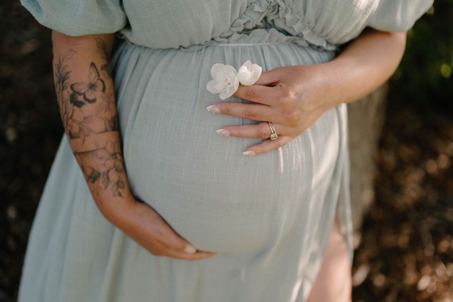Pregnant woman in sage green dress holding baby bump, showing tattoos and ring, holding white flowers outdoors.