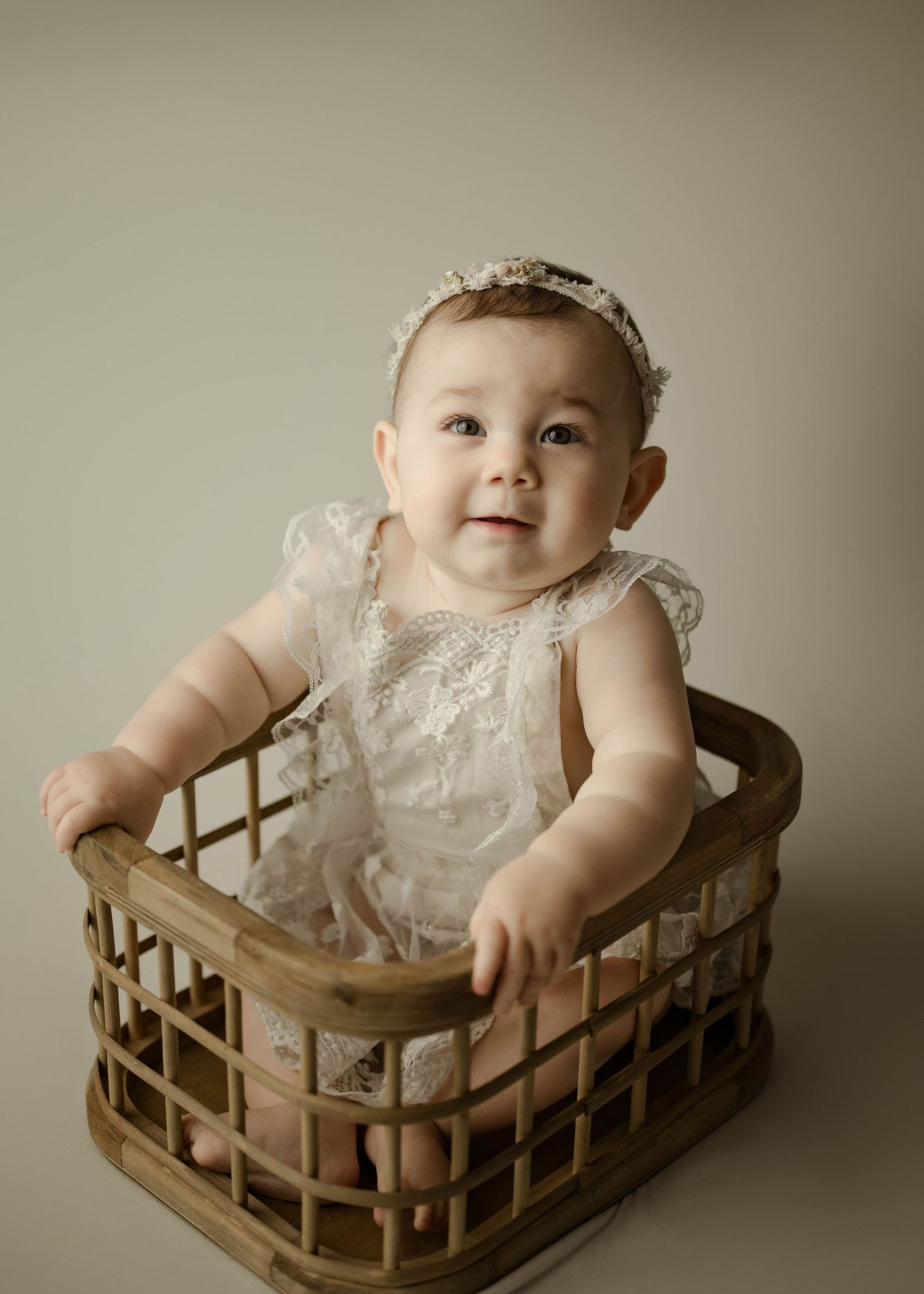 Smiling baby girl wearing lace romper and headband sitting inside a wooden wicker basket prop.