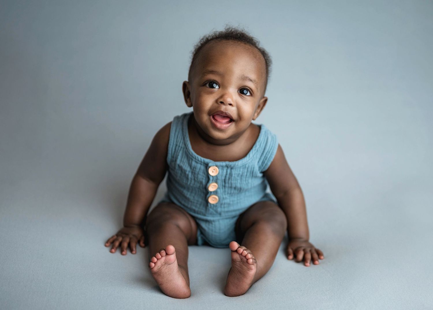 Smiling baby in blue sleeveless romper sitting on gray background.