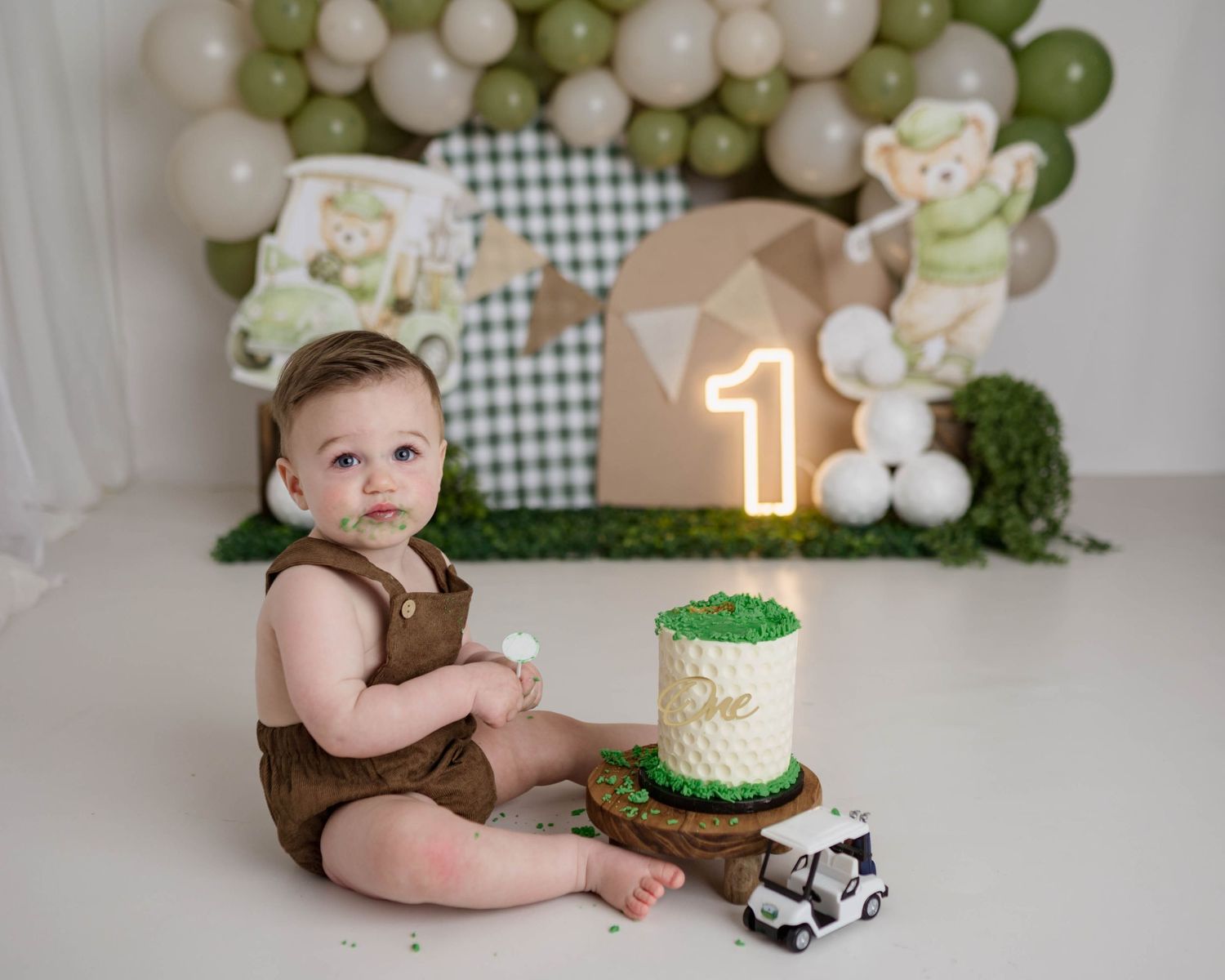 Baby's first birthday cake smash photo with green golf theme, balloon backdrop, number one candle, and adorable brown romper outfit.