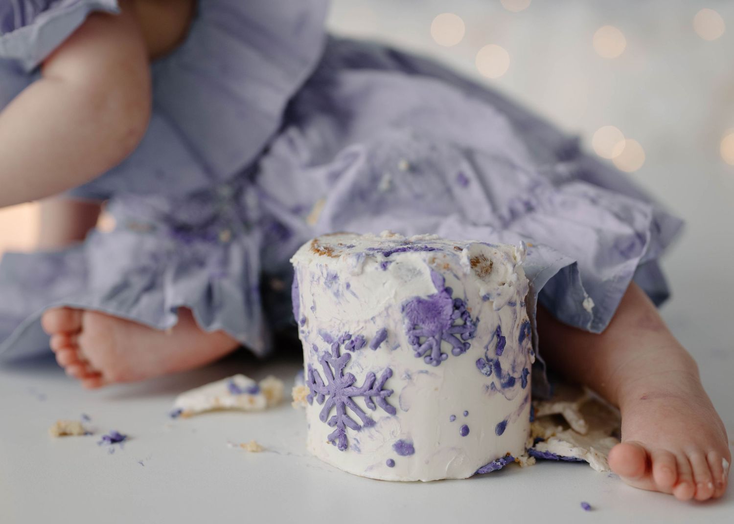 Close-up of baby feet beside a smashed white cake decorated with purple snowflakes on white floor.