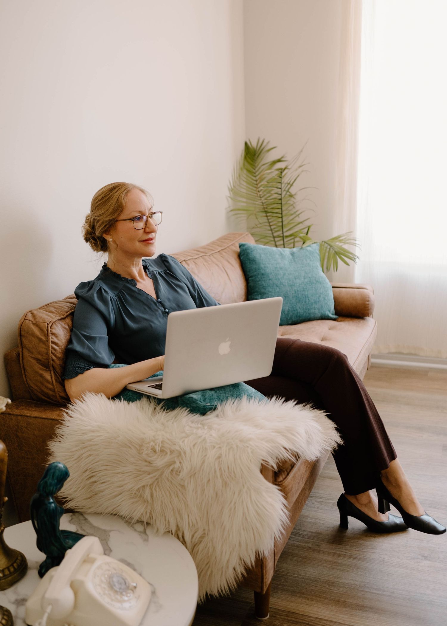 Woman in teal blouse working on MacBook laptop while sitting on brown leather sofa with fur throw.