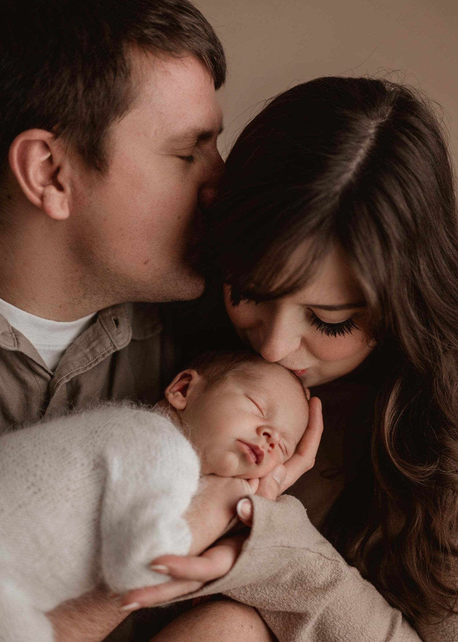 New parents sharing a tender kiss on their sleeping newborn baby wrapped in white.