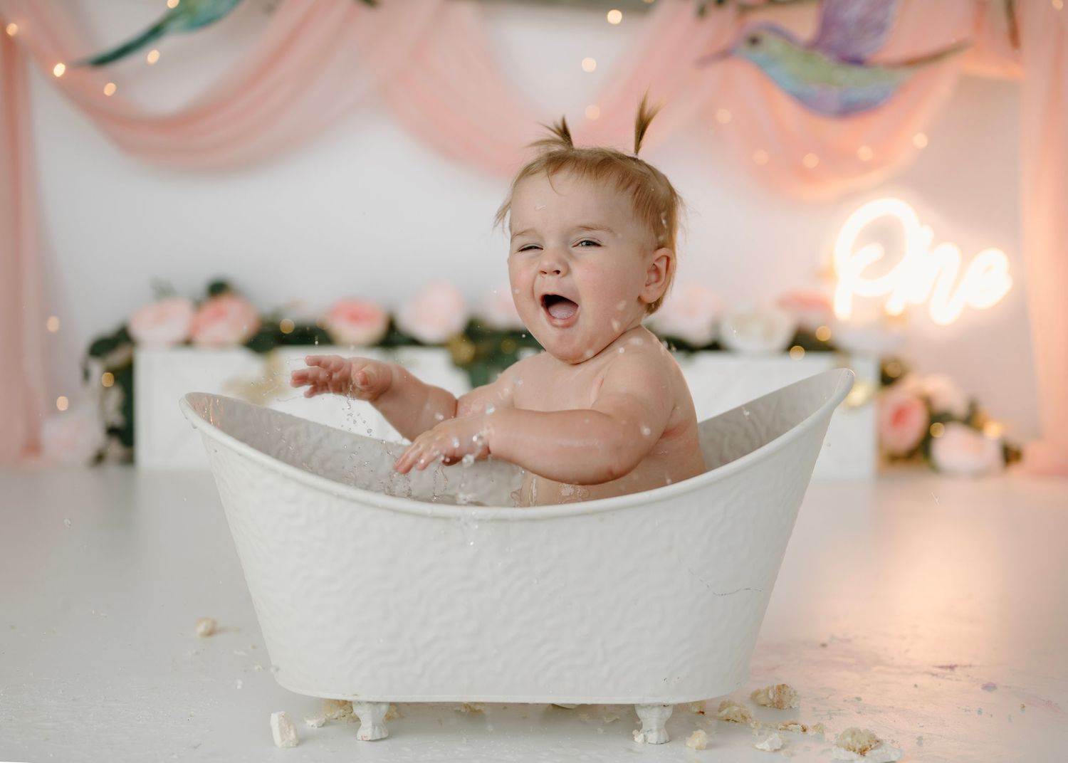 Laughing baby girl sits in a white clawfoot bathtub during a floral hummingbird-themed birthday bath session.
