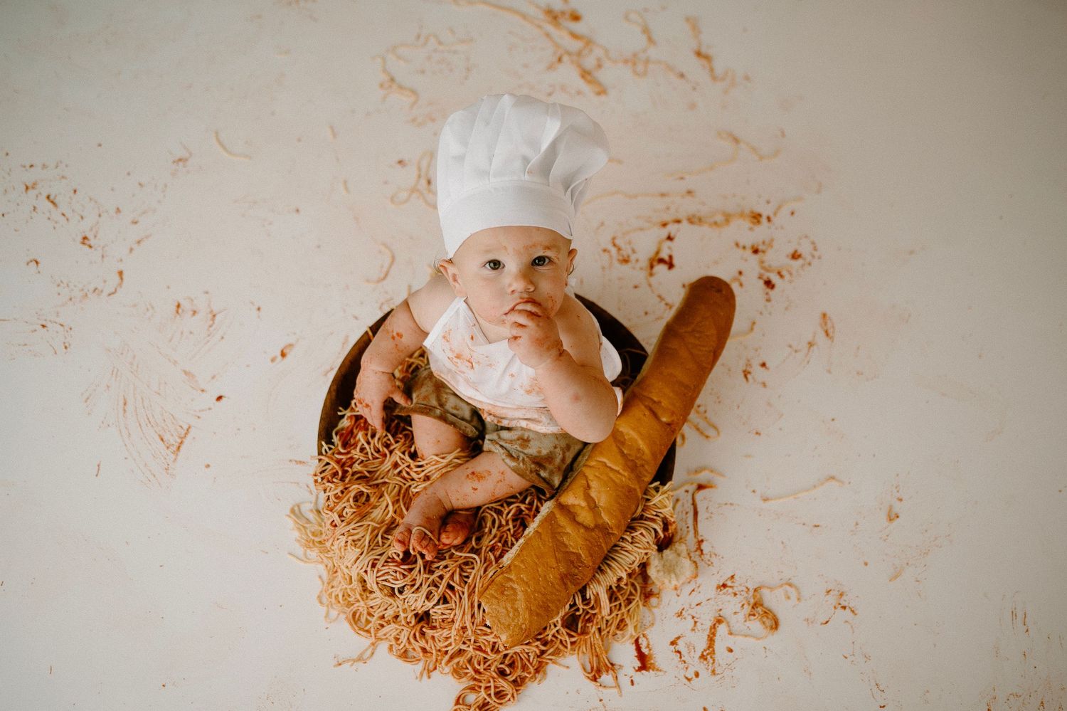 Overhead view of baby in chef hat lying in spaghetti bowl with baguette at birthday smash session.