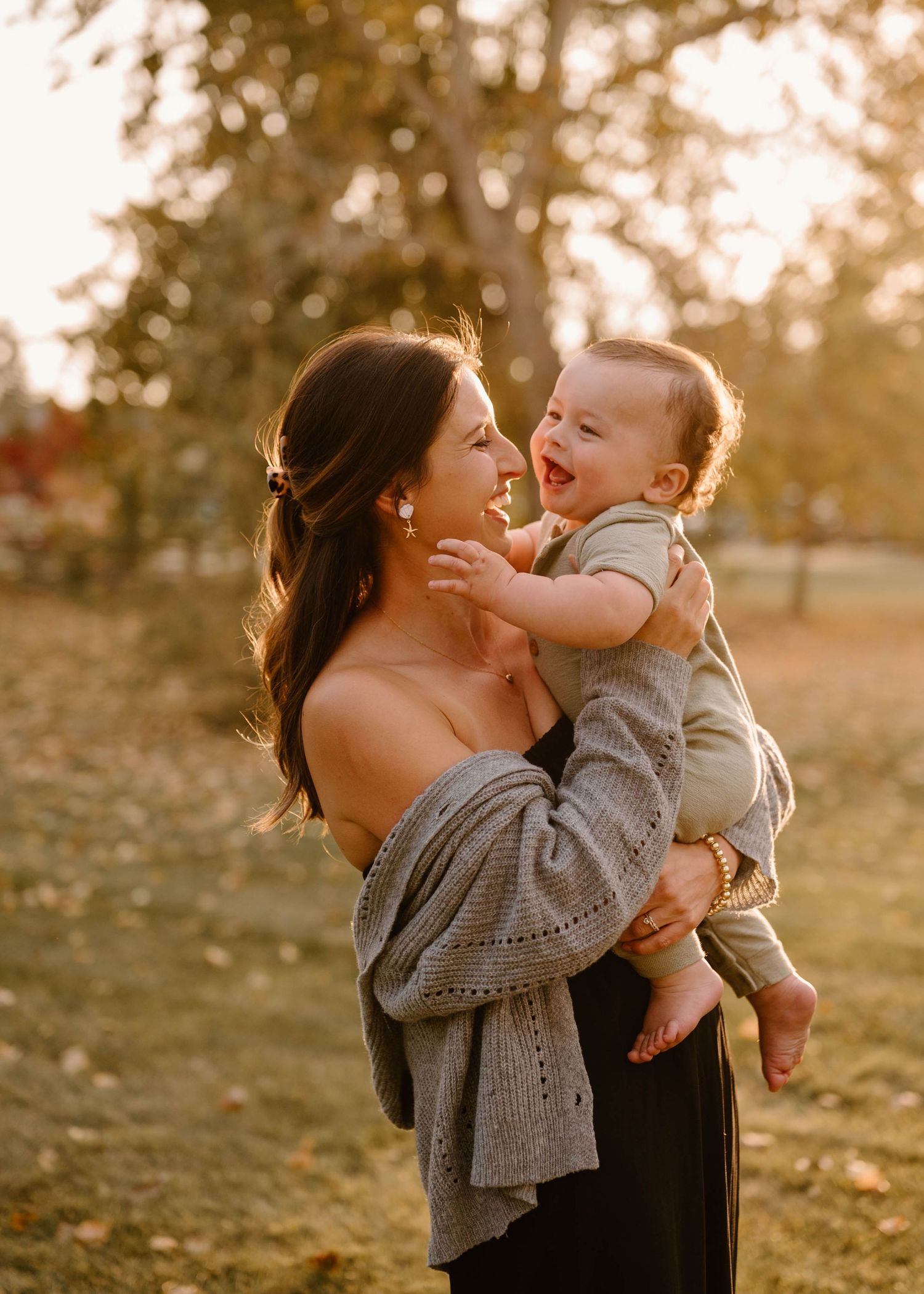 Joyful mother lifting laughing baby in sunlit autumn park, sharing a playful golden hour moment.