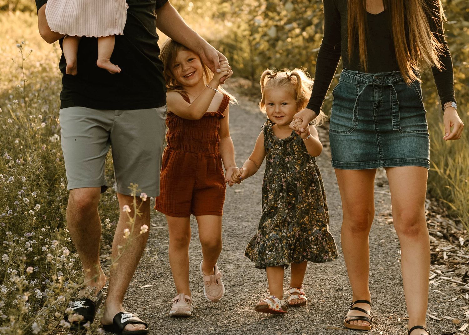 Family of four walking hand in hand on a sunny path through wildflowers.