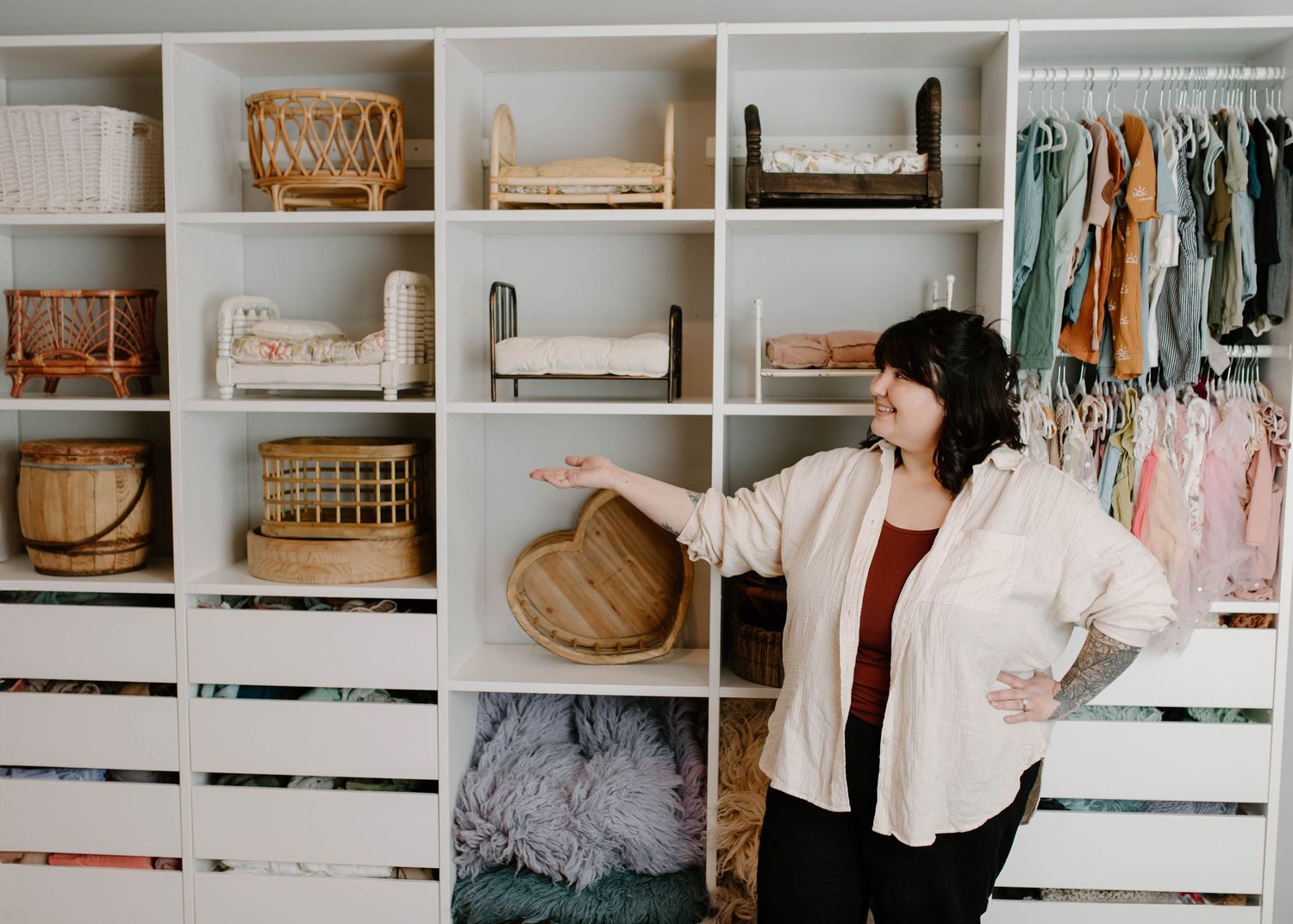 Woman organizing a stylish white shelving unit with baskets, decor, and hanging children's clothing in a bright room.