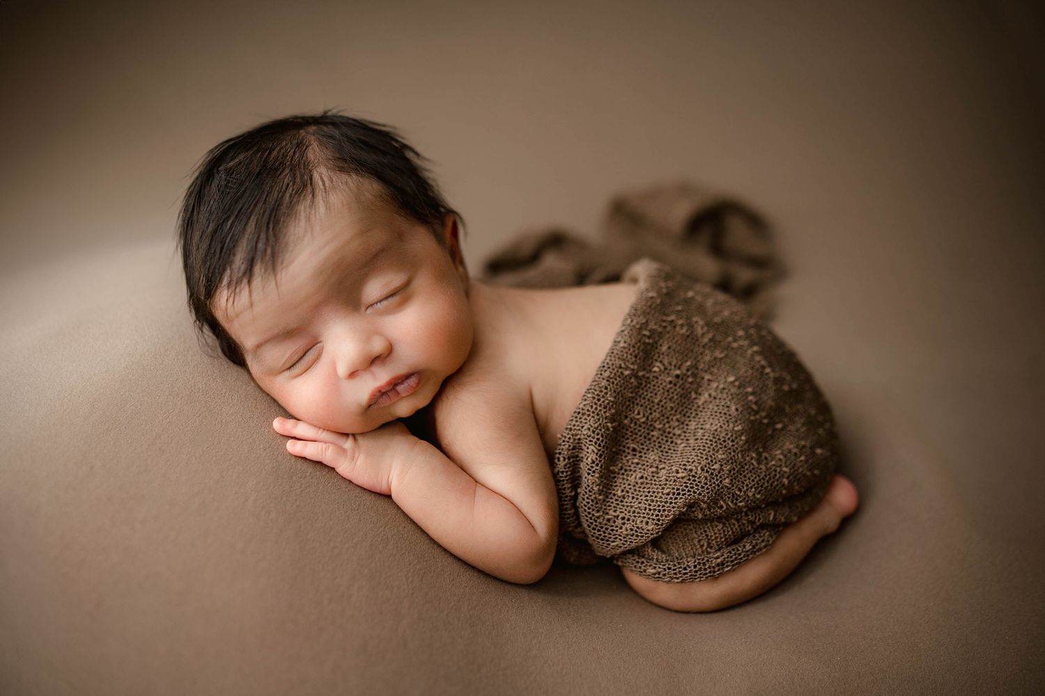 Peaceful newborn baby sleeping curled up on brown backdrop wrapped in soft brown fabric.
