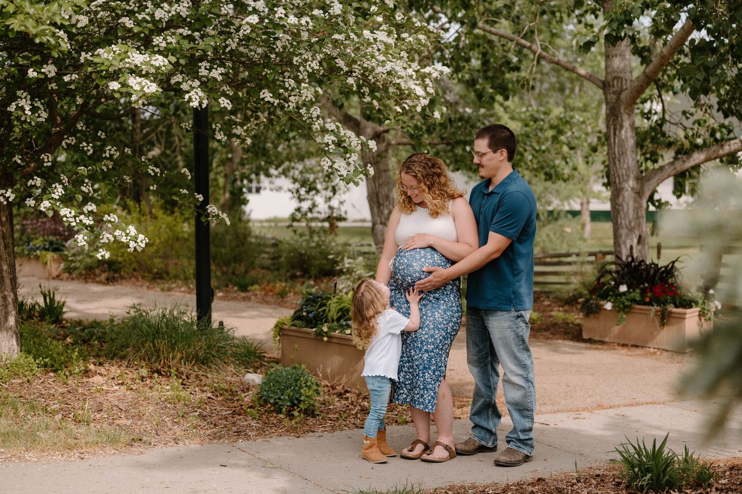 Pregnant woman with family outdoors in garden setting, husband embracing her belly while child stands nearby.