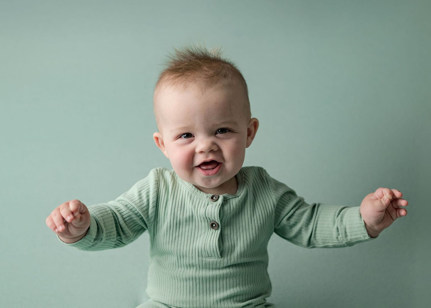 Happy laughing baby in mint green ribbed romper sitting with arms outstretched during studio milestone session.