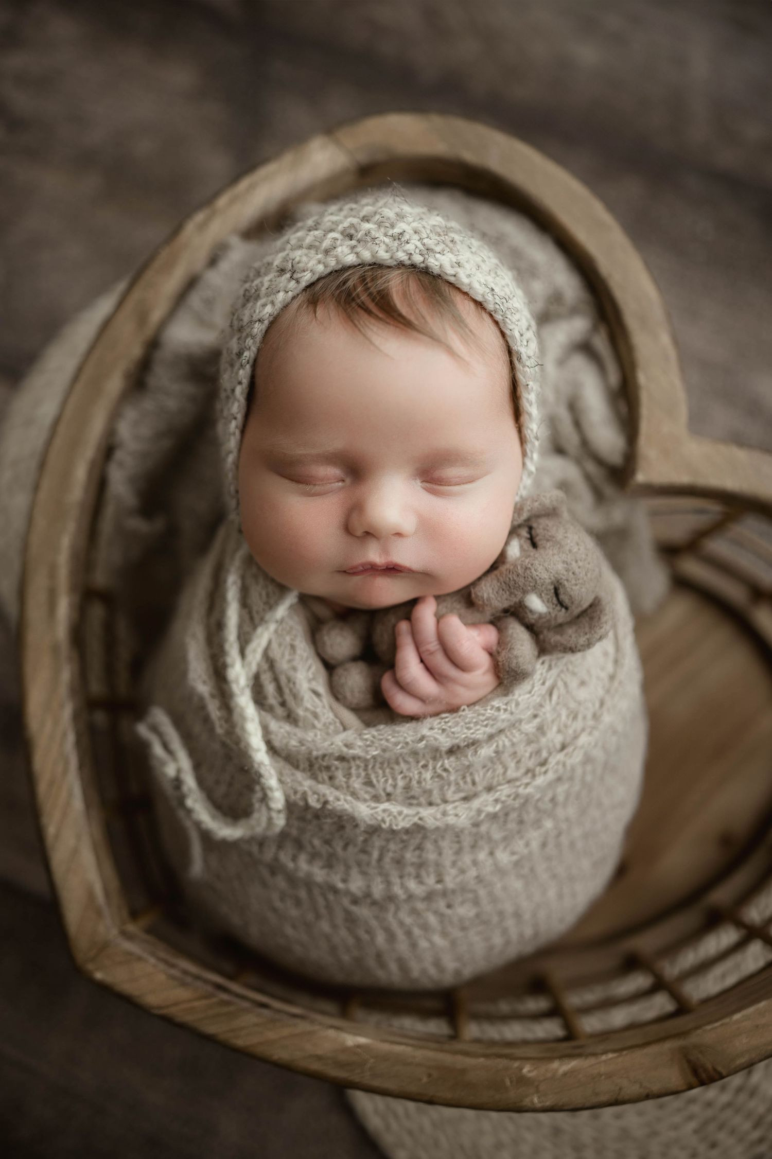 Sleeping newborn baby swaddled in beige knit wrap wearing bonnet, posed in rustic wooden heart-shaped basket.